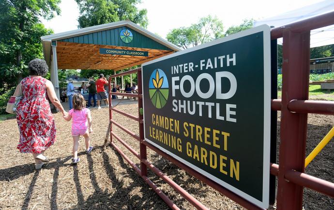 In the foreground, the backs of a mother and daughter holding hands and walking next to a gate with sign stating: Inter-Faith Food Shuttle. In the background, covered structure with sign that says: Community Classroom, with people gathered.