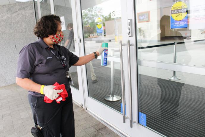 A white female RCC staff member with short wavy hair in gray short-sleeved uniform polo and dark-colored pants spraying a cleaning product on the glass doors of the Main Entrance with her left hand while holding a red cloth in her right hand.